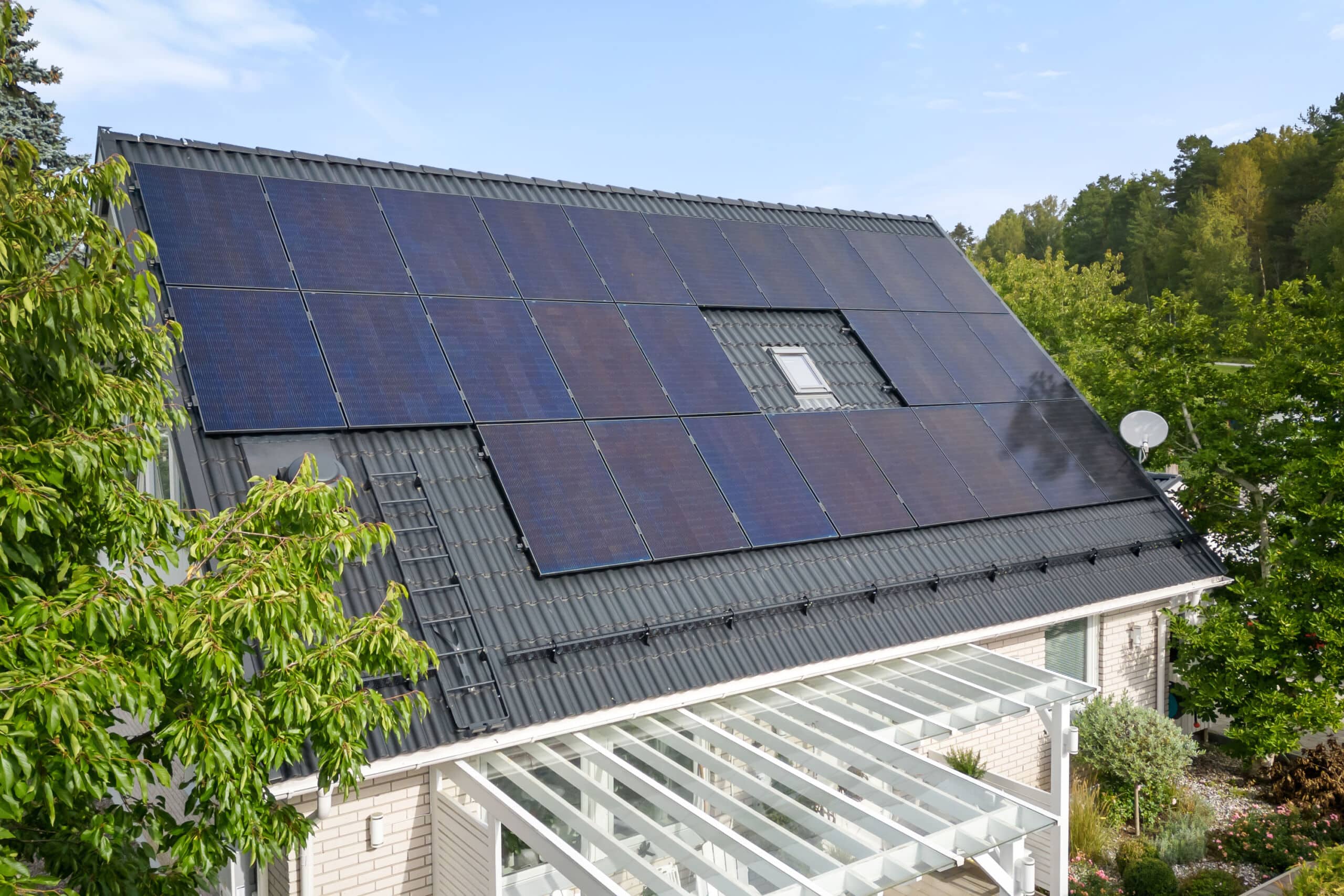 Svarta solceller på villa, klarbli himmel och fin pergola feamförhuset. Grön häck i förgrunden.