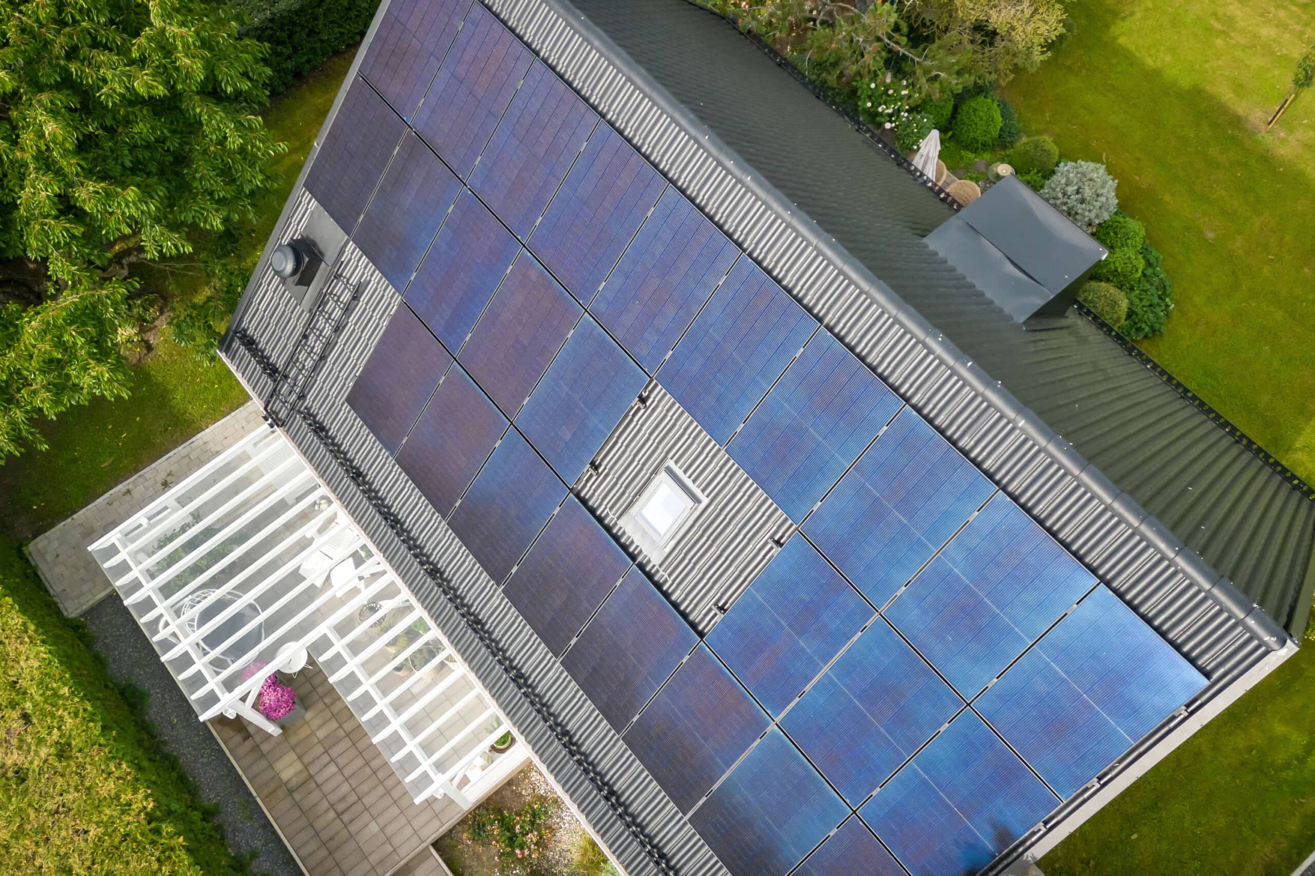 Svarta solceller på villa, klarbli himmel och fin pergola feamförhuset. Grön häck i förgrunden.