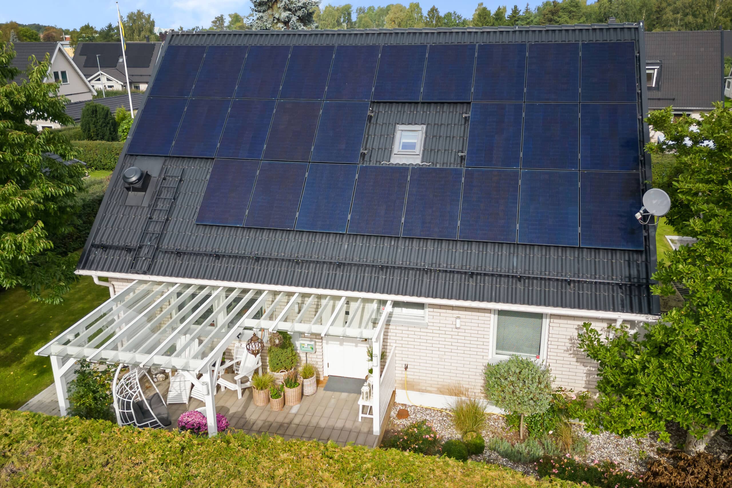 Svarta solceller på villa, klarbli himmel och fin pergola feamförhuset. Grön häck i förgrunden.