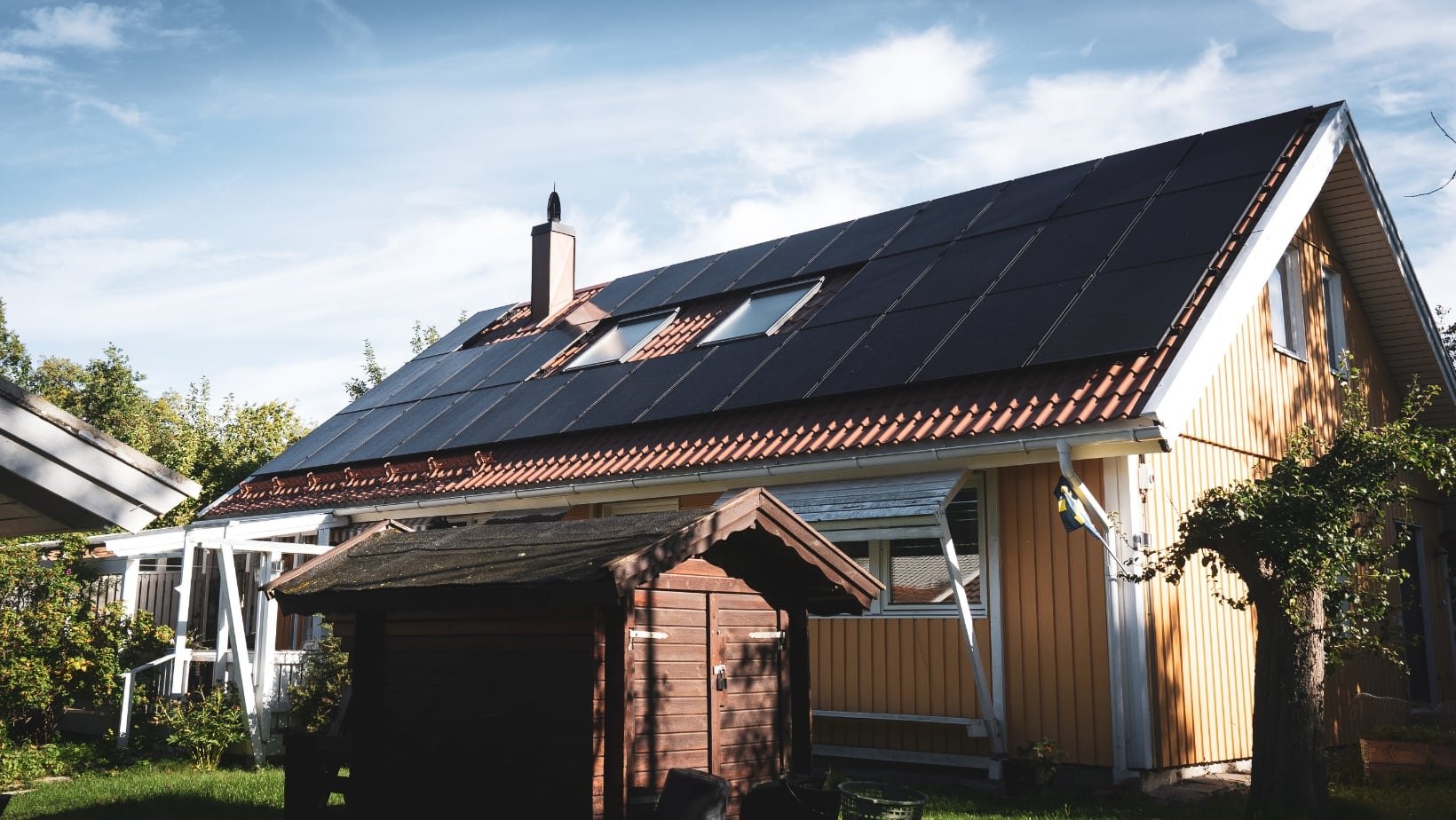 Svarta solceller som producerar grön el på gul villas tak, blå himmel med slöjmoln i bakgrunden och äppelträd och lillstuga i förgrunden.