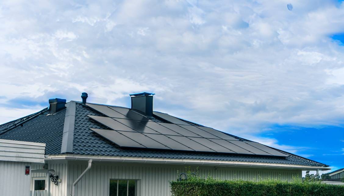 Vit villa med solceller på svart tak. Grön häck i förgrunden och blå himmel med slöjmoln i bakgrunden